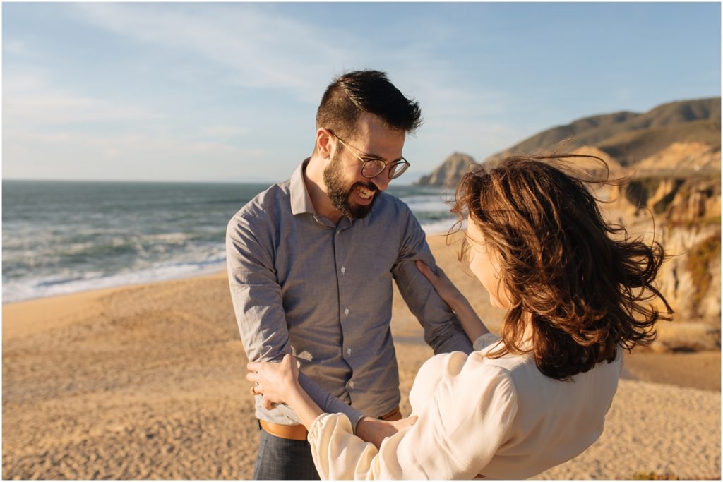 Couple embraces and laughs together during their Half Moon Bay engagement photos with an epic view behind them
