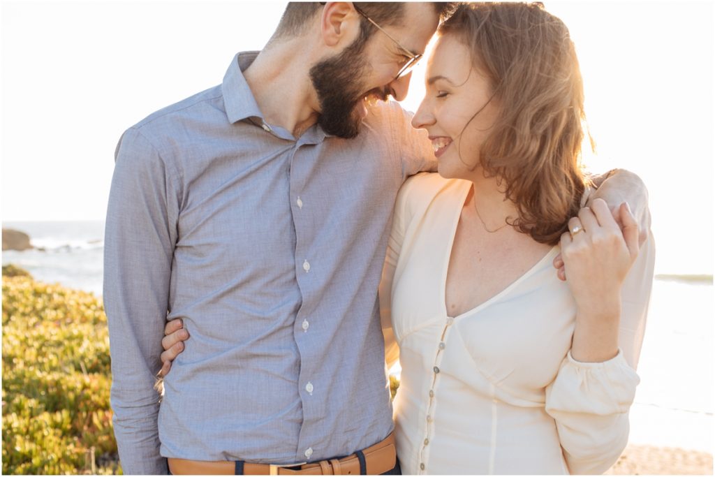 Man kisses woman behind the hear while they smile during their Half Moon Bay engagement photos