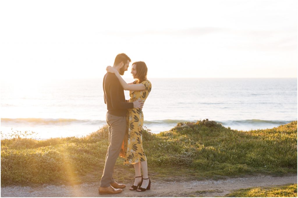 Man and woman embrace during their California engagement photos on a cliff over the ocean in the sunshine