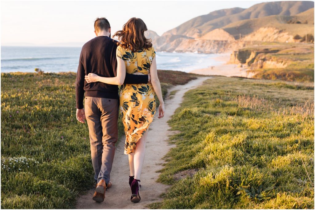 Man and woman walk arm in arm during their Half Moon Bay engagement photos on a cliff over the ocean in the sunshine