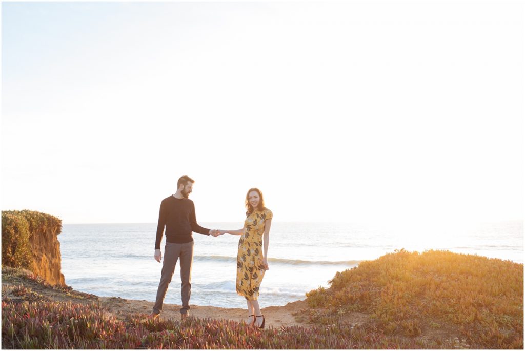 Man and woman hold hands during their Half Moon Bay engagement photos on a cliff over the ocean in the sunshine