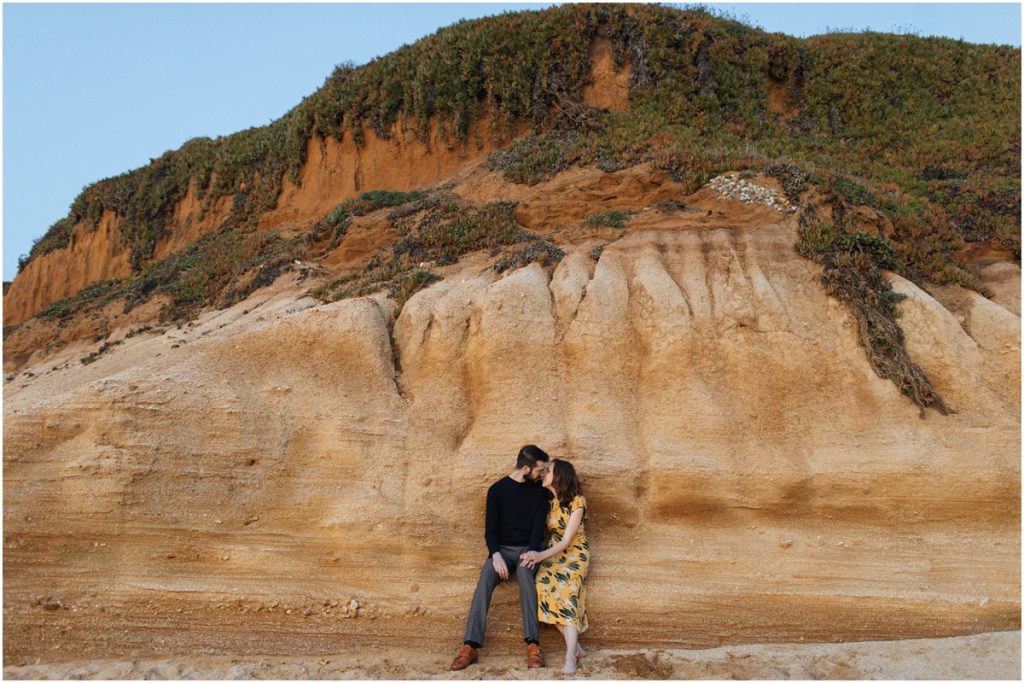 Couple sits together and kisses on the red cliffs during their Half Moon Bay engagement photos