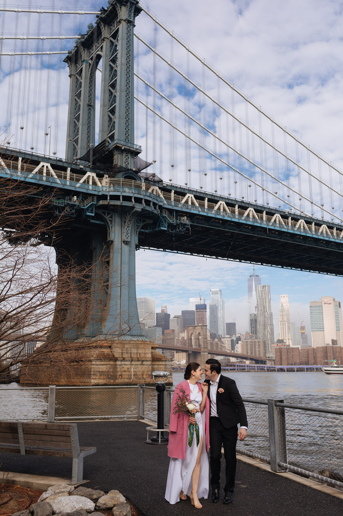Bride and groom stand smiling in front of the Manhattan Bridge during their morning Brooklyn elopement