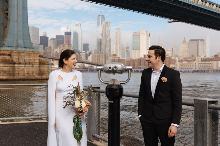 Bride and groom stand next to binoculars during their Brooklyn elopement with the NYC skyline behind them