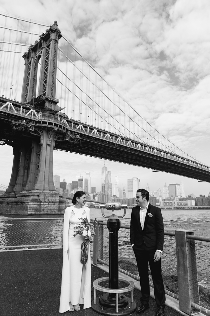 Bride and groom stand next to binoculars during their Brooklyn elopement with the NYC skyline behind them in black and white