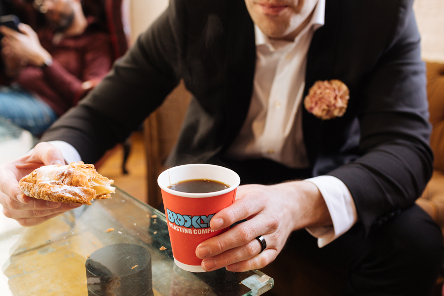 Groom drinks a cup of coffee and eats a pastry from the Brooklyn Roasting Company after their sunrise Brooklyn elopement