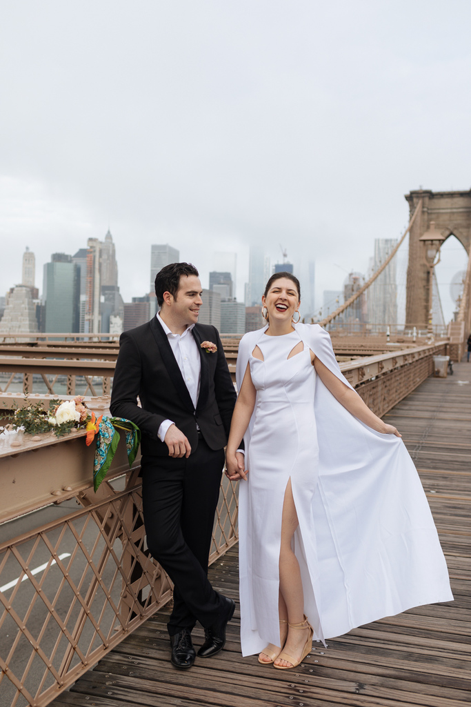 Bride and groom laugh together on the Brooklyn Bridge after their Brooklyn elopement: Groom looks on while bride shows off wedding dress cape