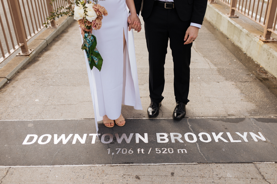 The bride and groom stand over a sign that says how far away is Brooklyn after their Brooklyn elopement