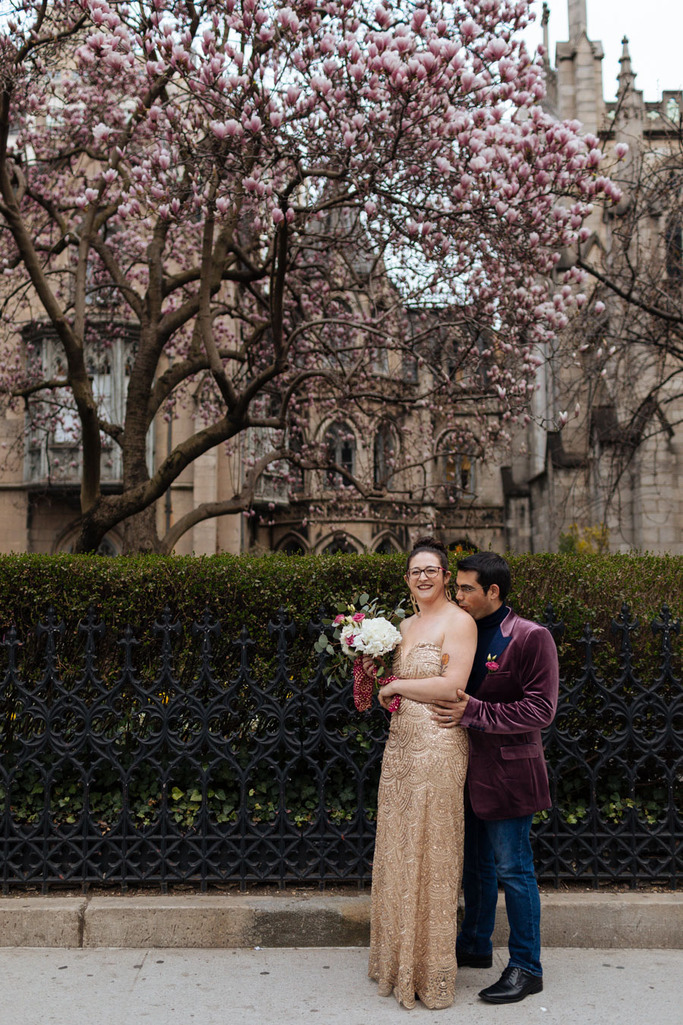 Groom kisses bride on the shoulder in front of the Grace Church under a large blooming magnolia tree during their East Village elopement in NYC