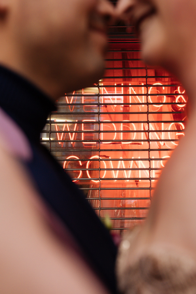 Bride and groom stand nose to nose in front of a neon sign that says Wedding during their East Village elopement in NYC