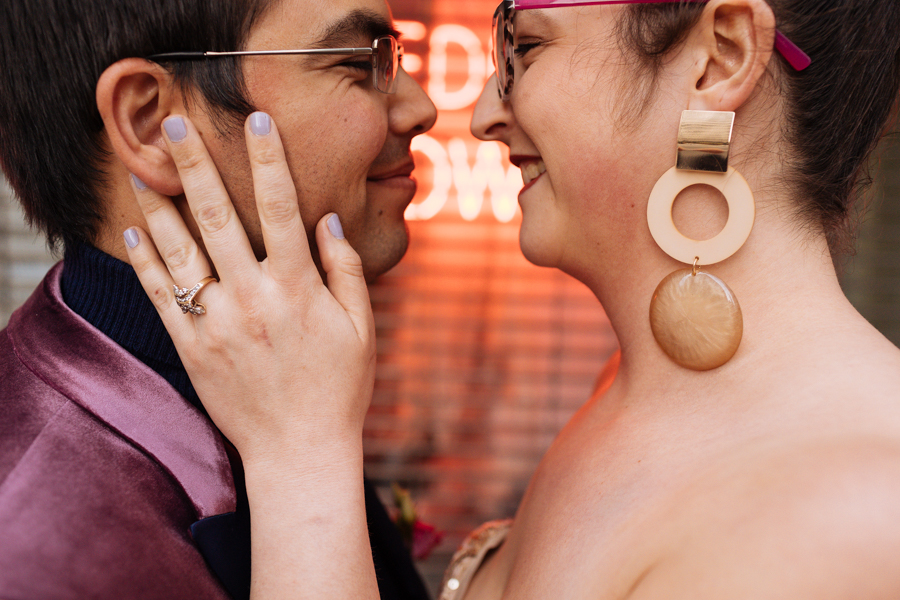 Bride and groom stand nose to nose in front of a neon sign that says Wedding during their East Village elopement in NYC