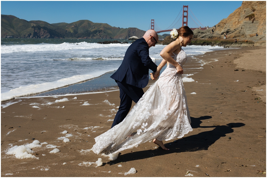 Bride and groom run away from the waves on the sand at Baker Beach in front of the Golden Gate Bridge during their San Francisco picnic wedding on the beach