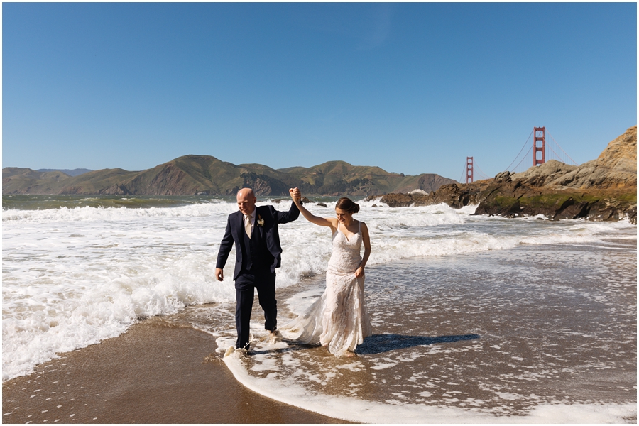 Bride and groom walk through the waves at Baker Beach hand in hand in front of the Golden Gate Bridge during their San Francisco picnic wedding on the beach