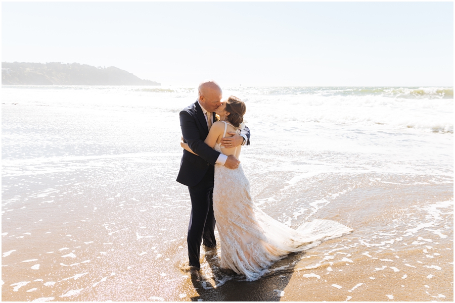 Bride and groom kiss in the waves at Baker Beach in front of the Golden Gate Bridge during their San Francisco picnic wedding on the beach